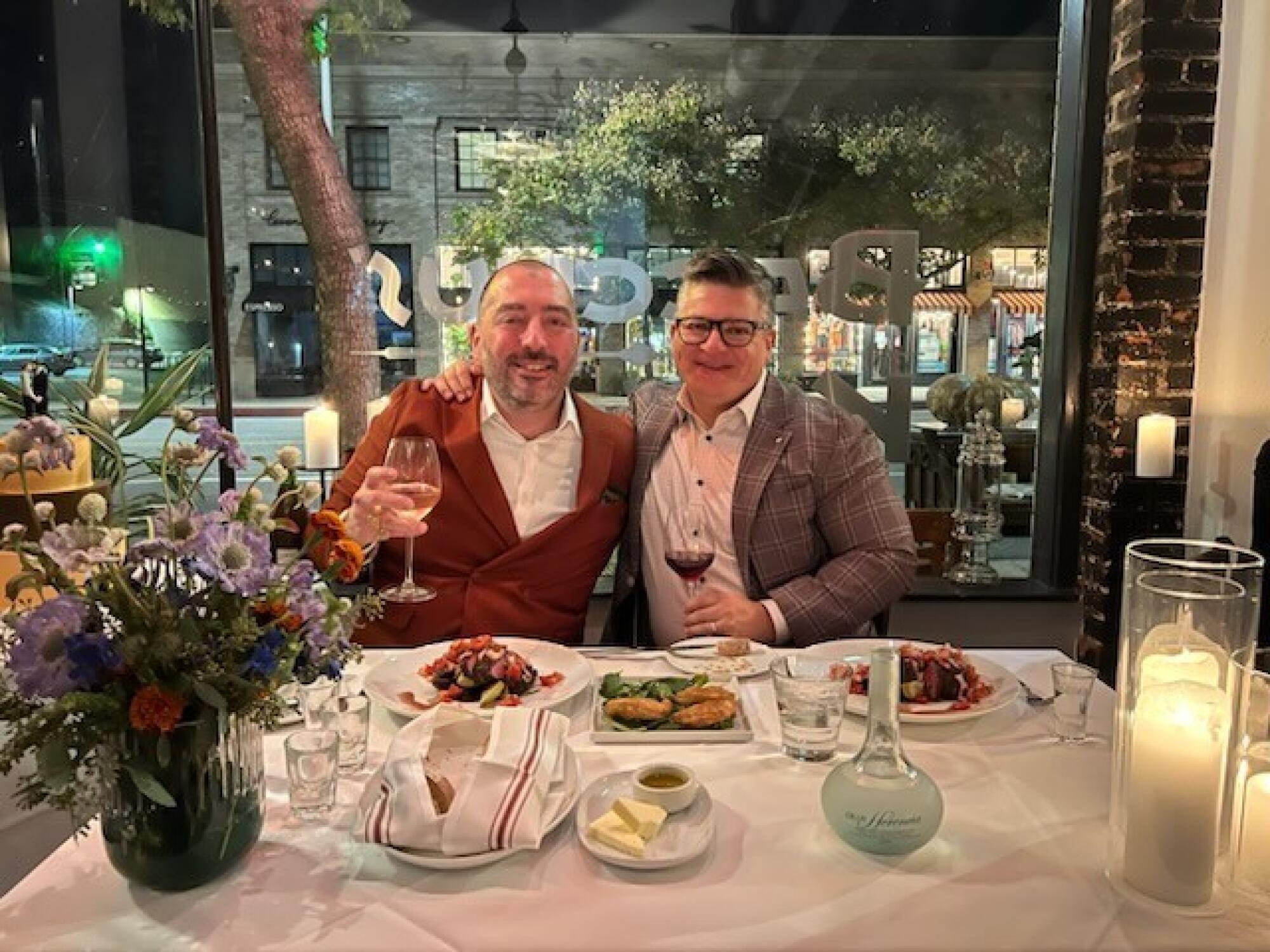 Two men in suits sit, arms around each other, with wineglasses in hand and food on the table before them.