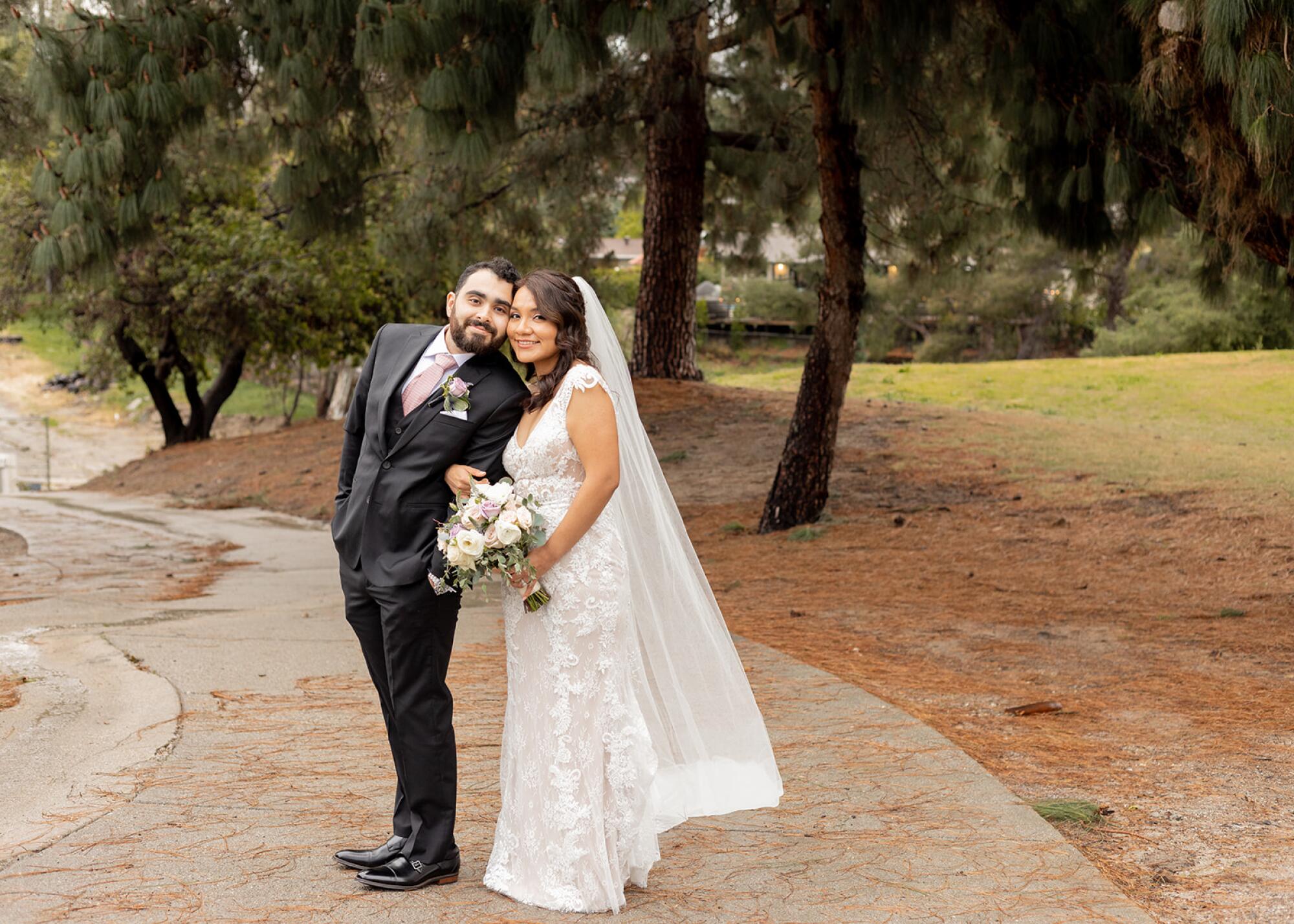 A man in a dark suit and a woman in a wedding dress with a bouquet stand outdoors in a park