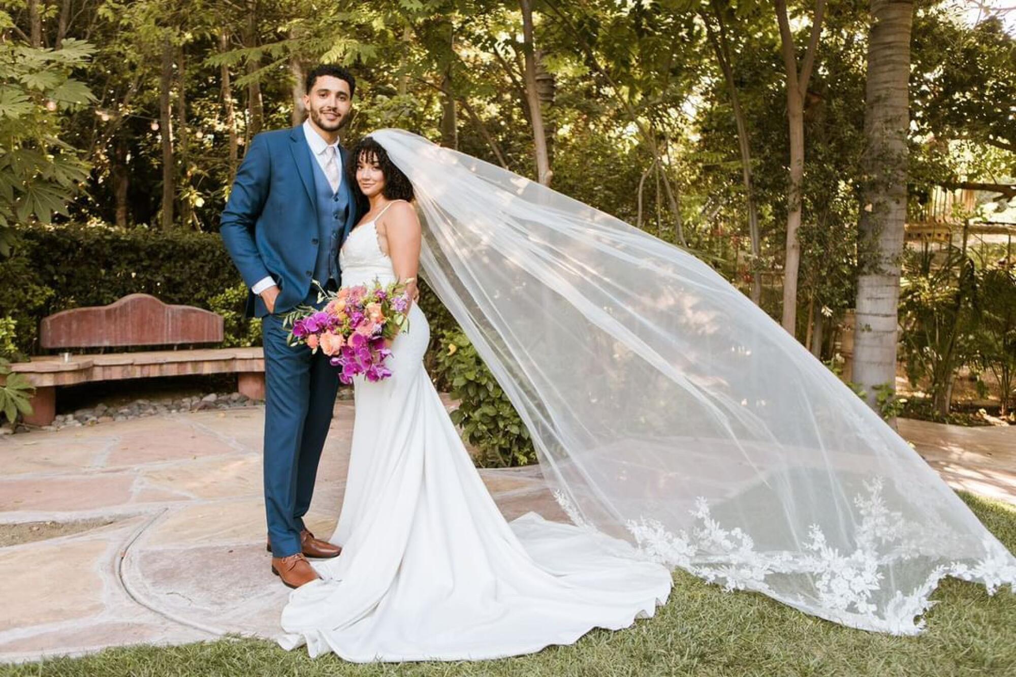 A tall man in a peacock-blue suit stands next to his bride, who has a long gown and veil and a pink bouquet