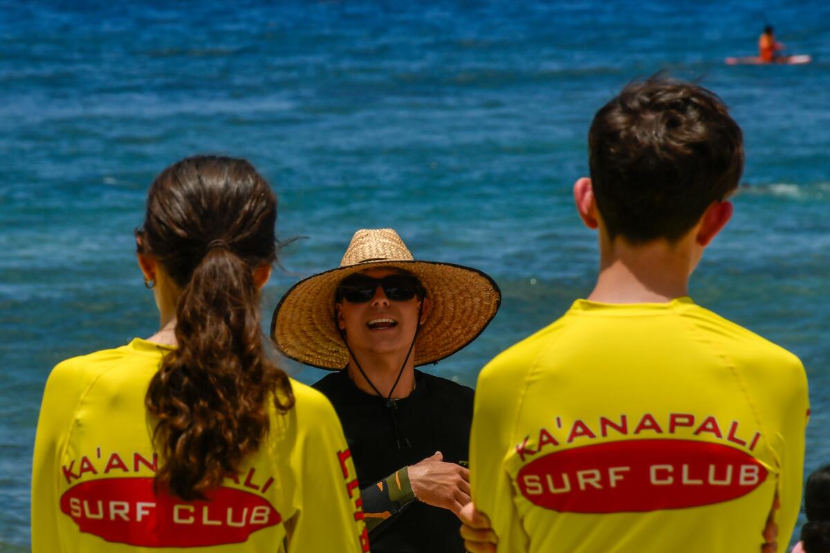 A person in a straw hat talks to two kids in Ka'anapali Surf Club T-shirts, the ocean behind them.