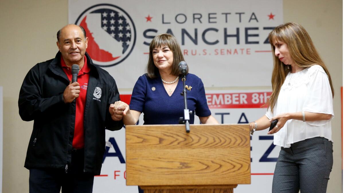 A woman dark hair, in a navy outfit, stands at a lectern, flanked by a man holding a microphone and a woman in a white top