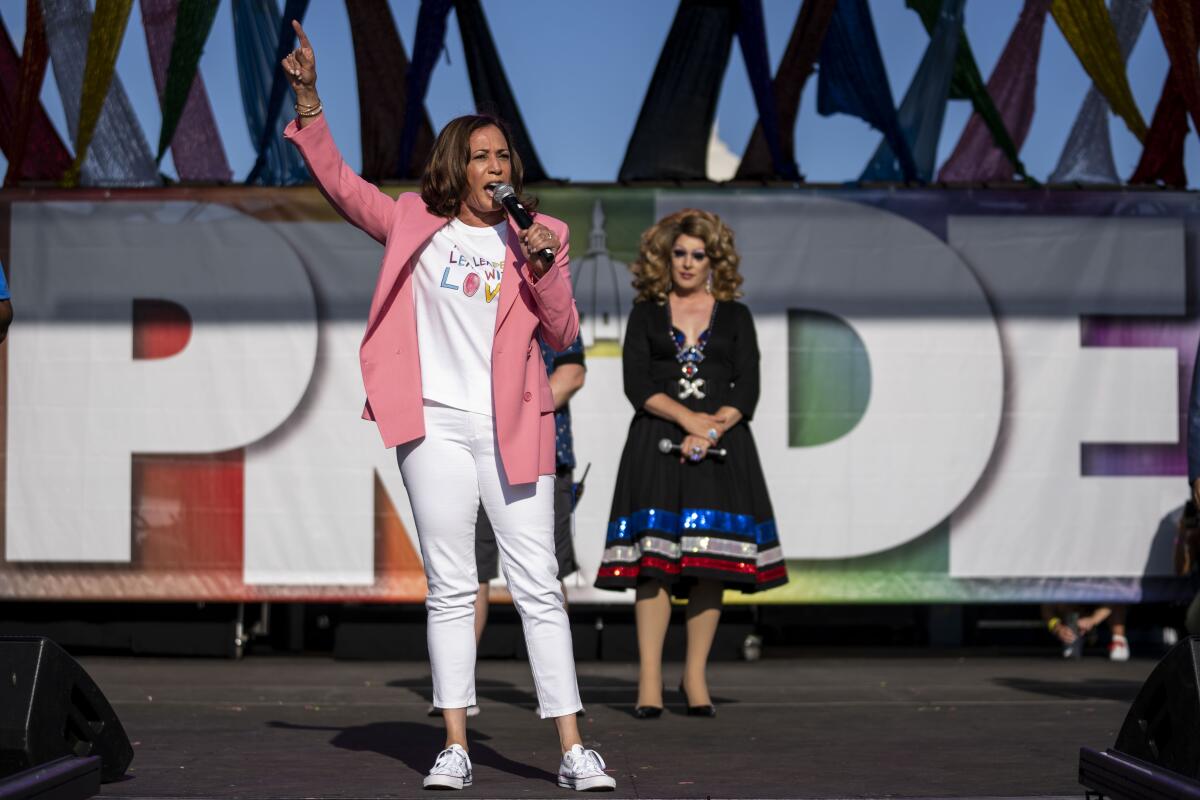 A woman in a pink jacket, white top and pants holds up a hand while speaking with a microphone