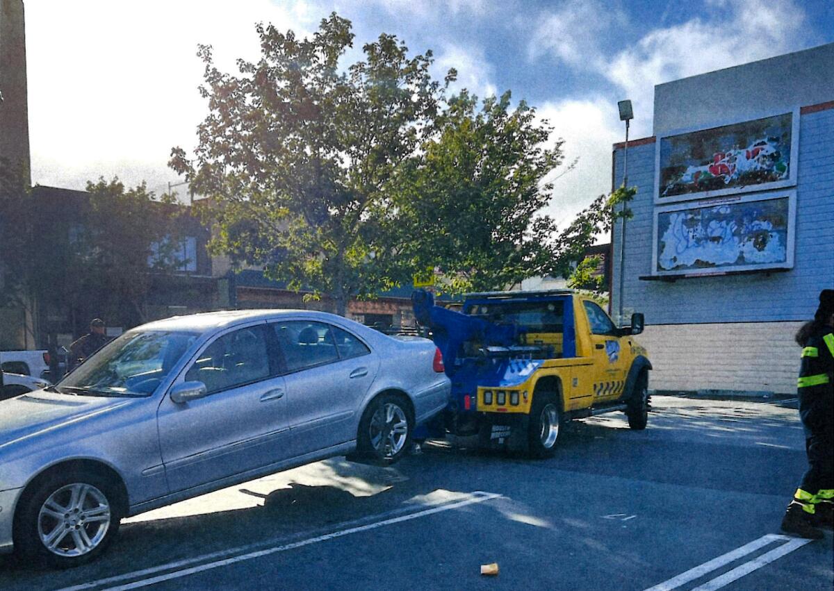 A yellow tow ruck pulls a silver car behind it near a building