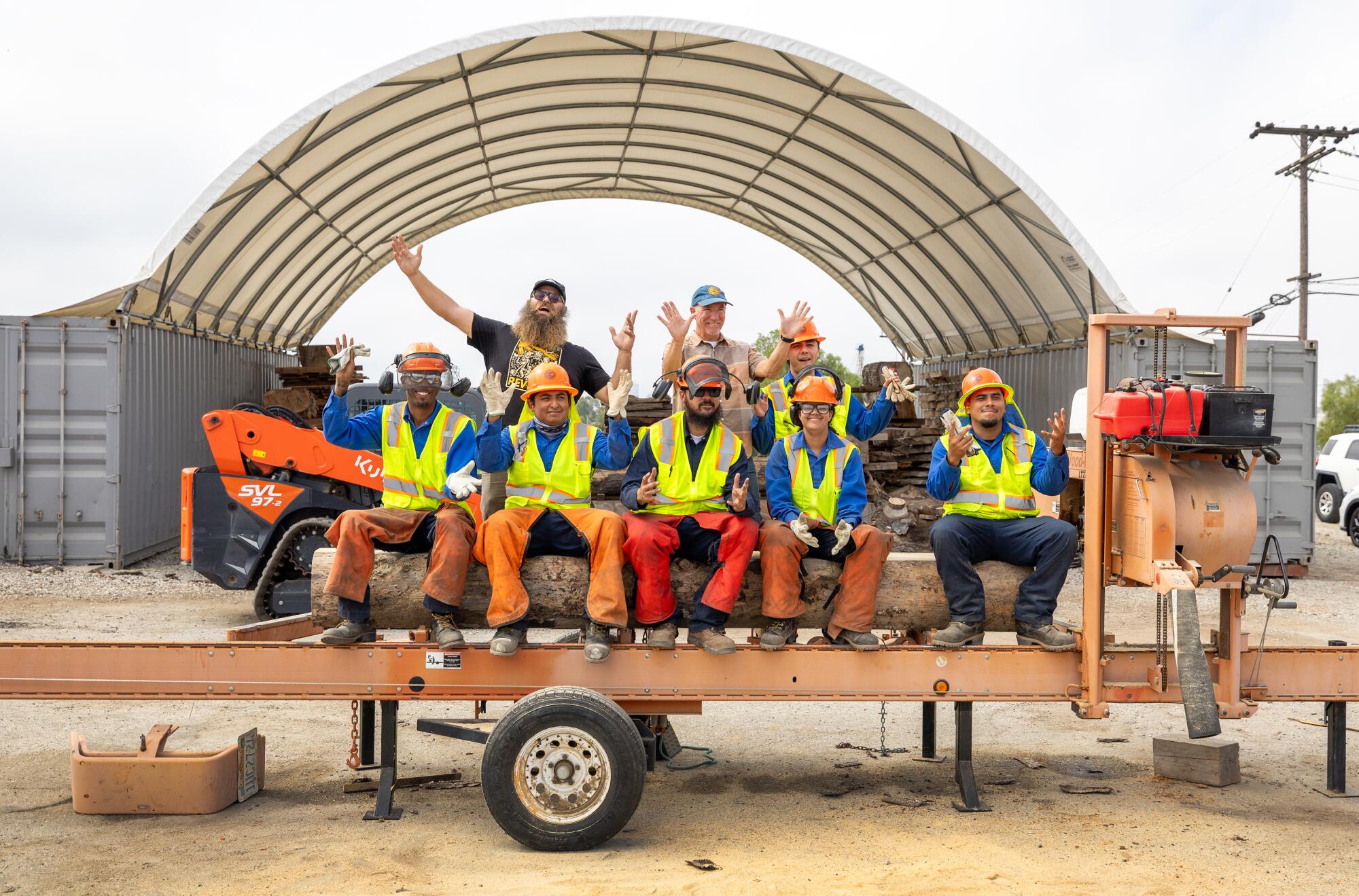 A group of people in orange hard hats and yellow reflective vests sit atop a log on a trailer and wave.