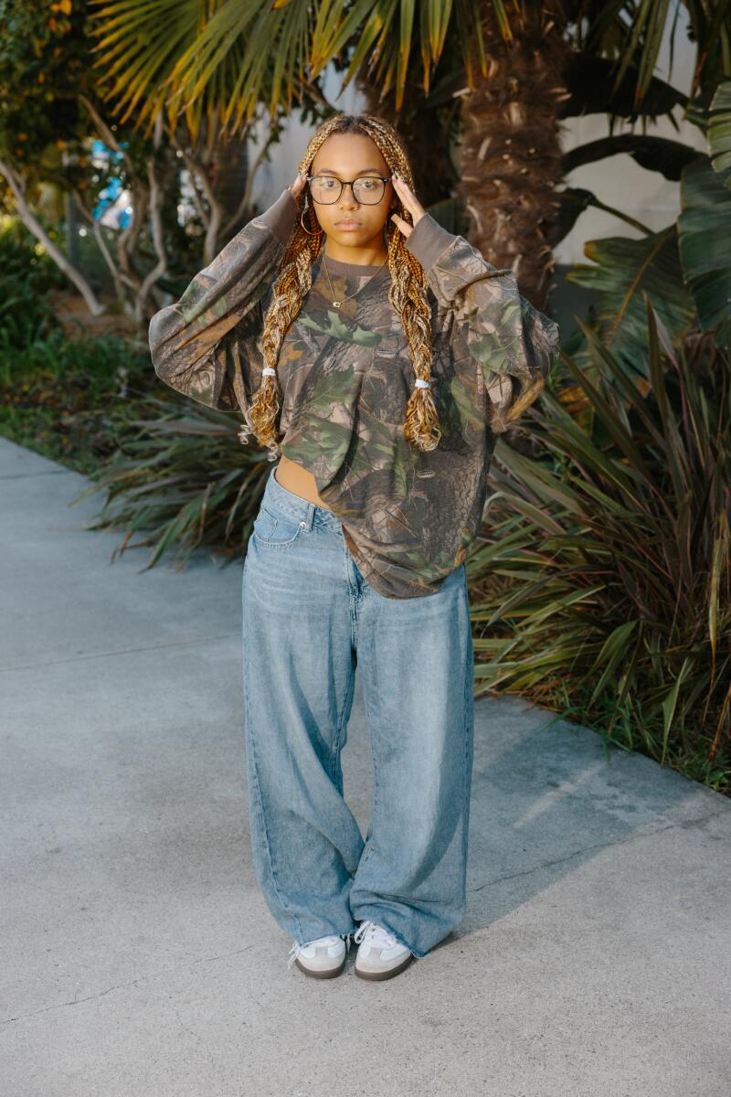 A girl posing for a portrait wearing glasses, a long-sleeved hunting camo shirt, baggy jeans and sneakers.