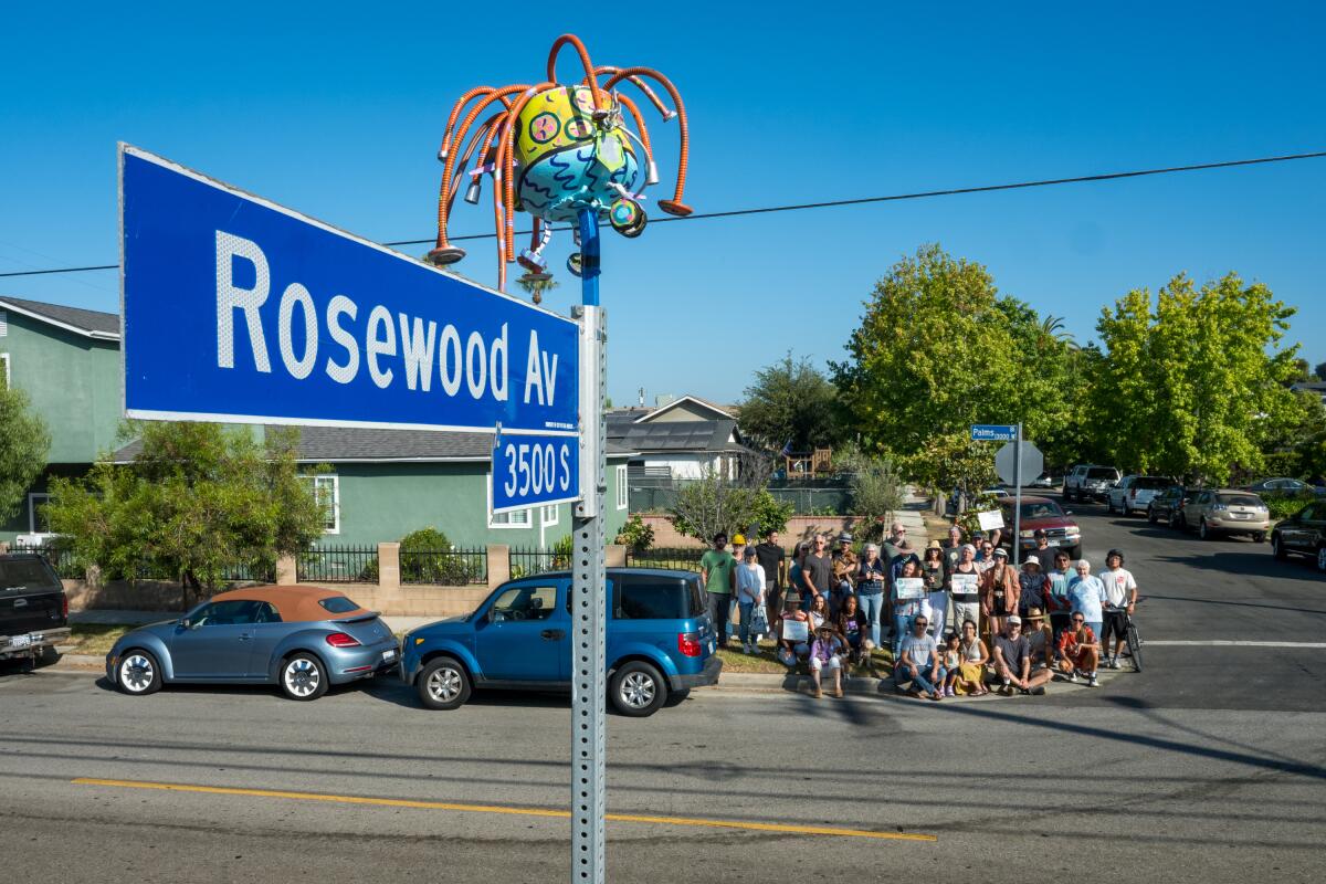 A streetsign topped with a sculpture and people gathered across the street 